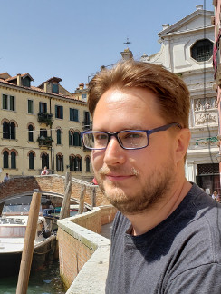 Picture of Lee. We see him standing outside in the sunlight. He is wearing glasses. In the background there are some buildings, a canal, small boat, and a small bridge. Maybe somewhere in Europe, like Venice?